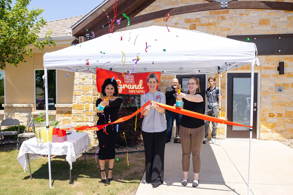 Three people from the massage therapy team Frisco cut a red ribbon under a canopy at a grand opening, with confetti in the air and a “Grand Opening” sign behind them, celebrating the launch of therapeutic massage Frisco TX.