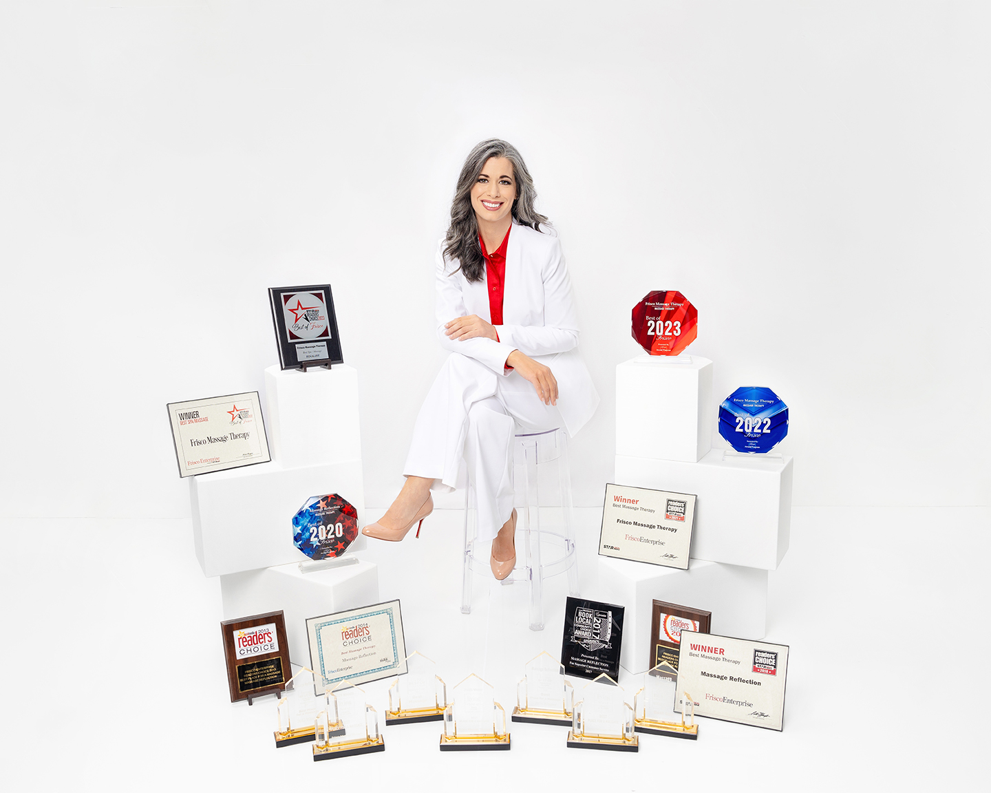 A woman in a white suit sits on a clear chair, surrounded by multiple awards for therapeutic massage Frisco, displayed on white stands and the floor, in a pristine white studio setting.