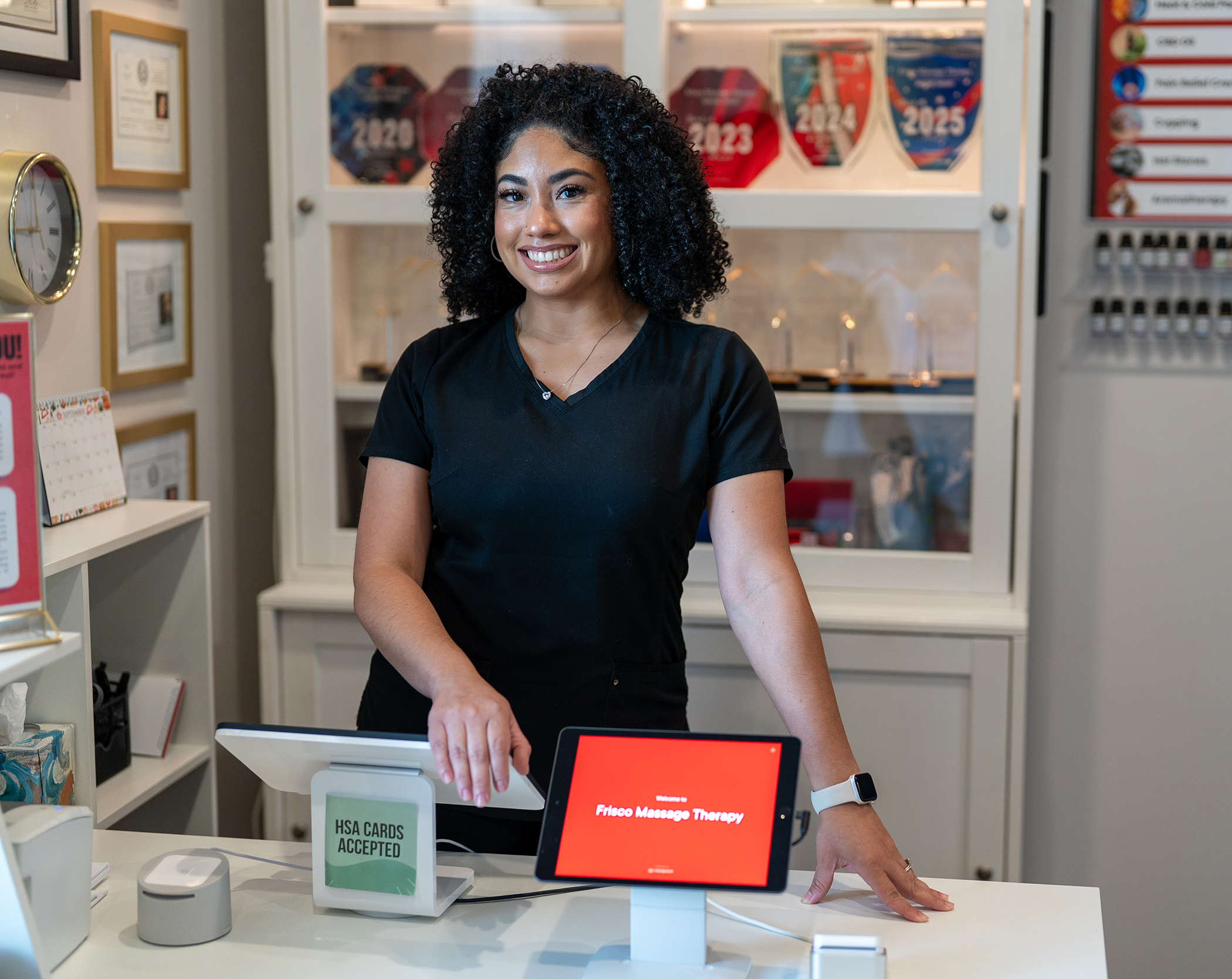 A woman stands behind a reception desk in a professional setting with a sign reading "Flores Massage Therapy" displayed on a tablet. Awards and certificates highlight expertise in MLD Frisco and lymphatic drainage in the background.