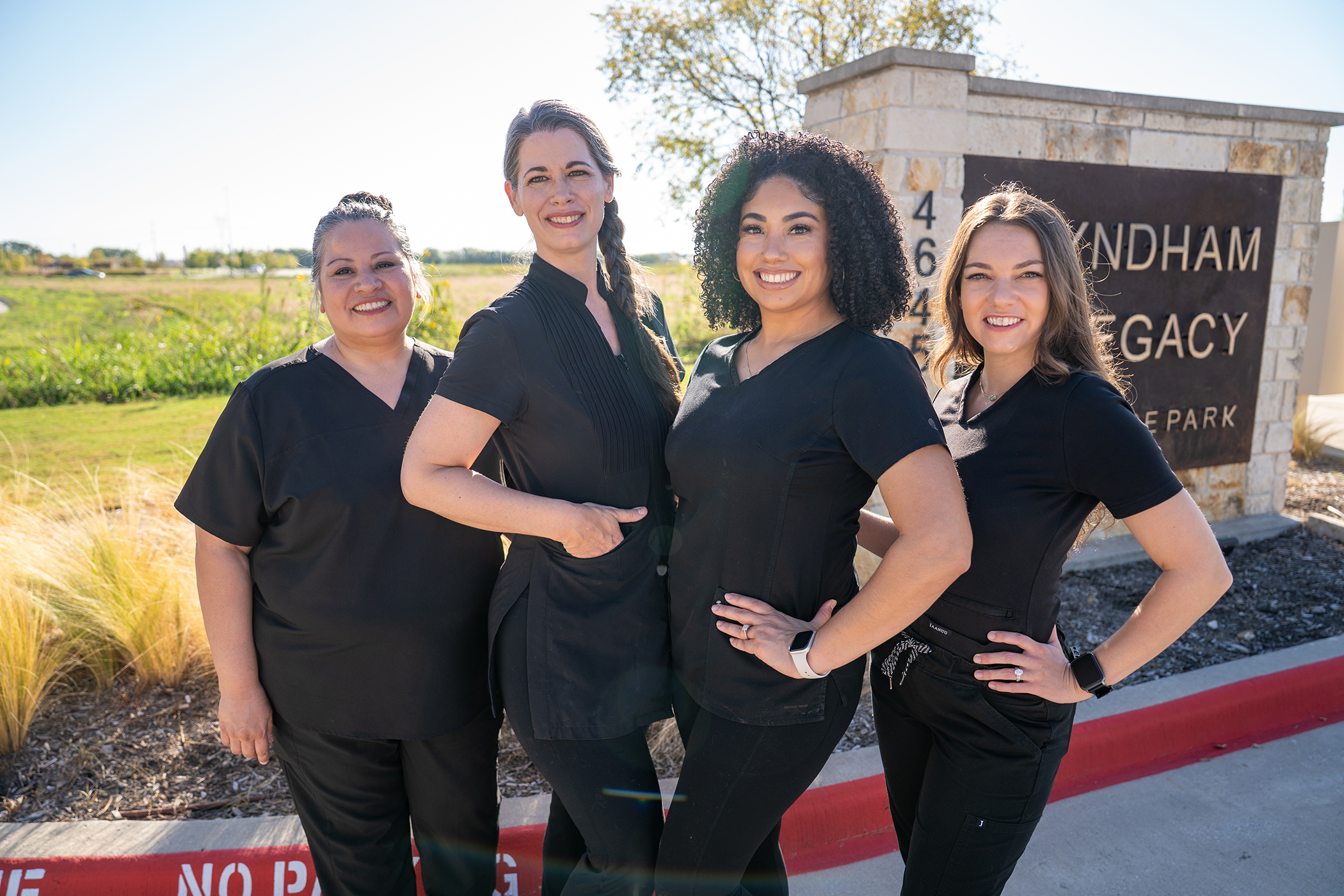 Four women in black uniforms stand smiling in front of a stone sign that reads "Wyndham Legacy Office Park" on a sunny day, offering expert pre-surgery lymphatic massage Frisco to support surgery recovery and well-being.