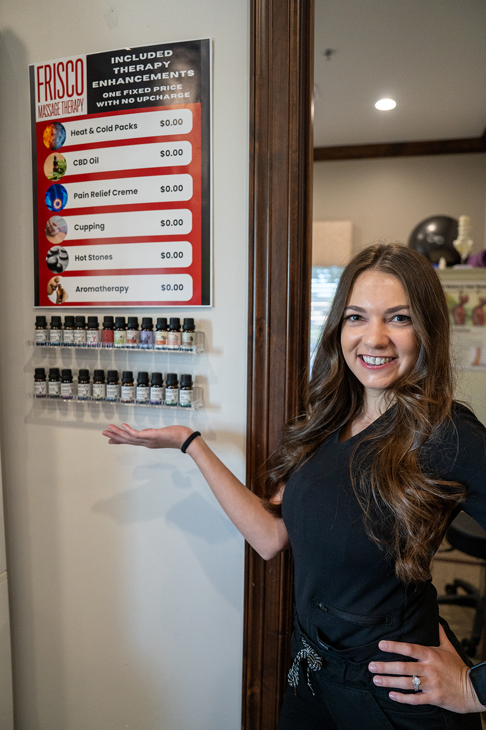 A woman in a black outfit stands indoors, smiling and gesturing toward a wall-mounted Therapeutic Massage in Frisco services and aromatherapy display.
