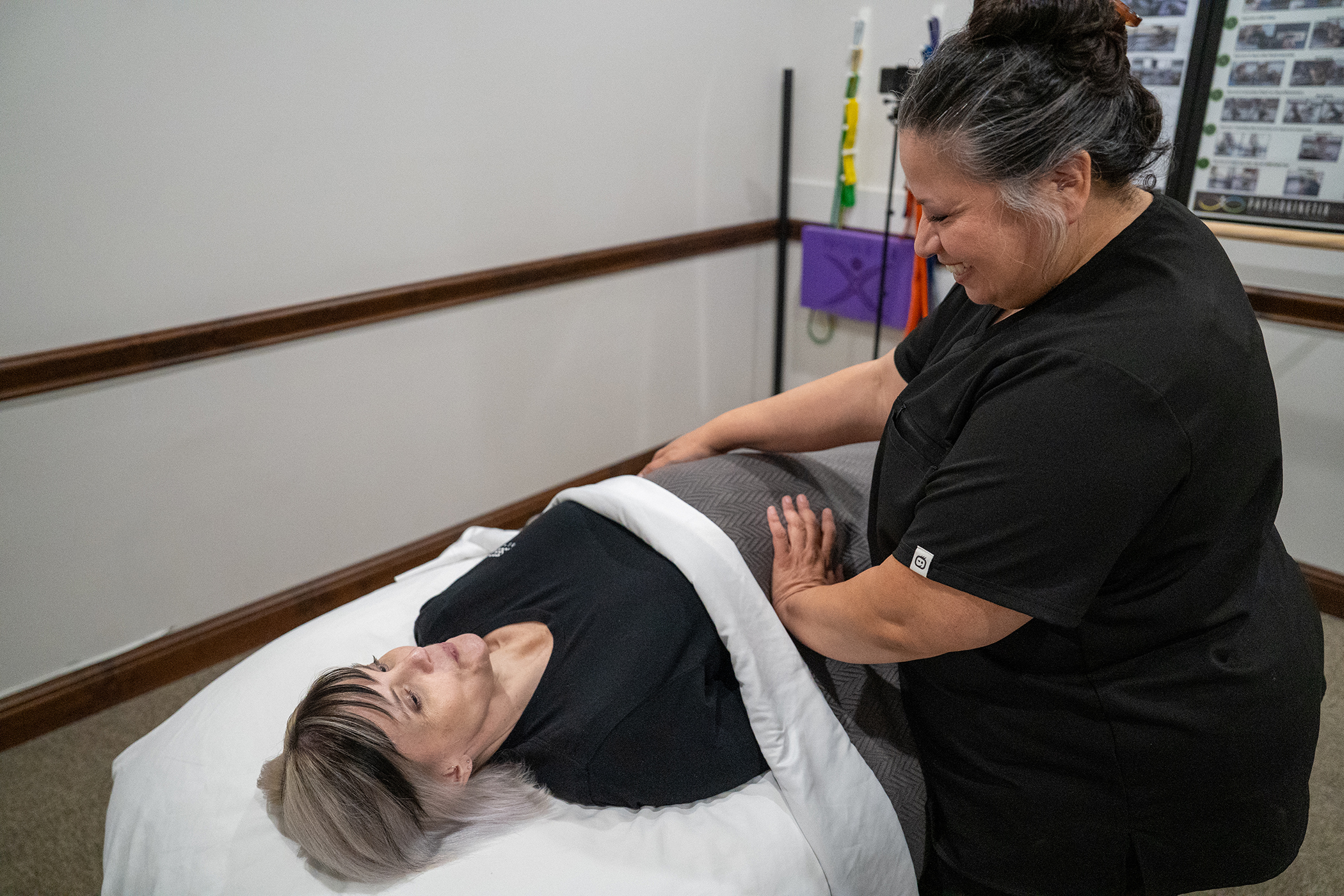 A woman lies on a treatment table covered with a blanket while a therapist in black scrubs stands beside her, smiling and providing expert prenatal massage Frisco care.