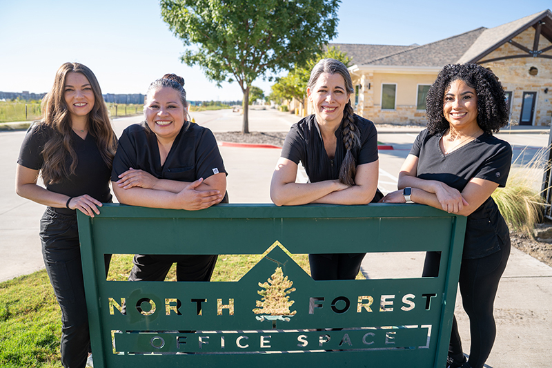 Four women in black uniforms stand and smile behind a green sign that reads "North Forest Office Space," ready to share their expertise in TMJ massage and injury recovery massage on a sunny day.