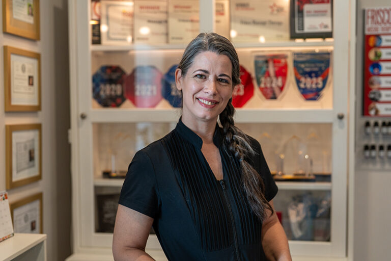 A woman with long gray hair in a braid stands smiling in front of a display of framed awards and certificates, celebrating her achievements in Prenatal Care and Frisco Massage Therapy.