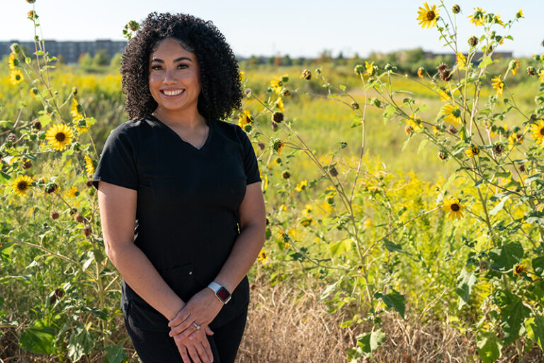 A woman with curly hair stands outdoors in a field of sunflowers, wearing black scrubs and a smartwatch, smiling at the camera—reflecting her dedication to oncology massage and cancer pain management.