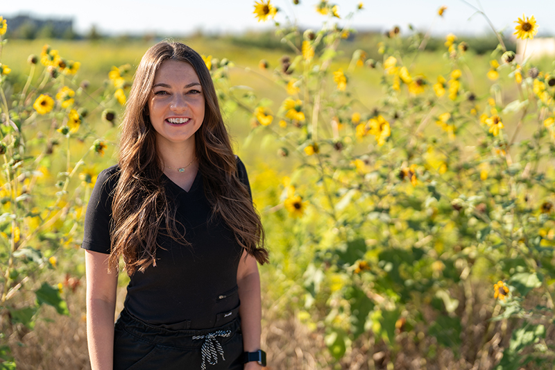 A woman with long brown hair wearing a black shirt stands smiling in front of tall sunflowers in a sunny outdoor field, reflecting the uplifting benefits of oncology massage for cancer pain management.