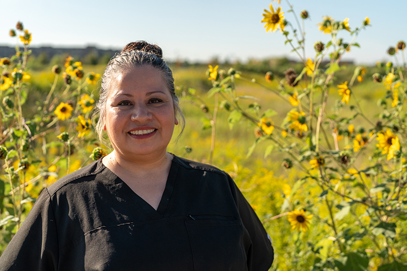 A woman in black scrubs stands smiling in a field of sunflowers under a clear sky, embodying the care and compassion found in oncology massage and cancer pain management.