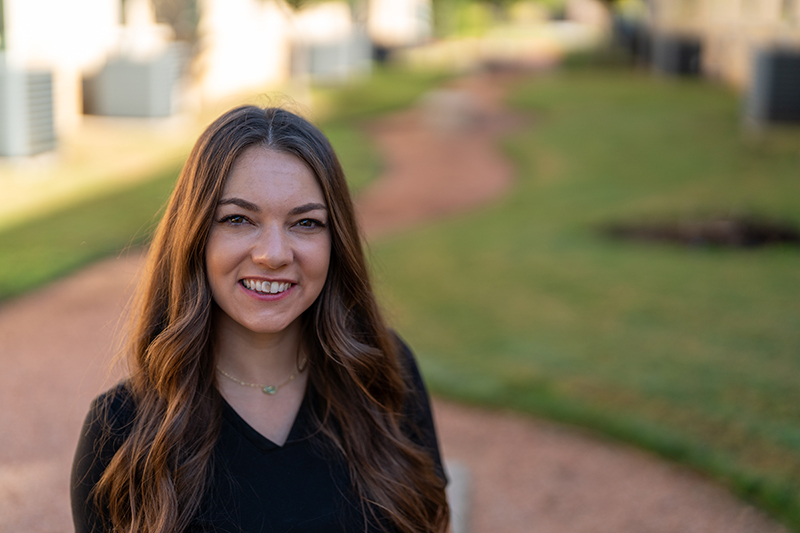 A woman with long brown hair and a black top stands outdoors on a gravel path, enjoying the fresh air—an ideal setting to relax and support her immune system boost inspired by lymphatic drainage massage techniques.