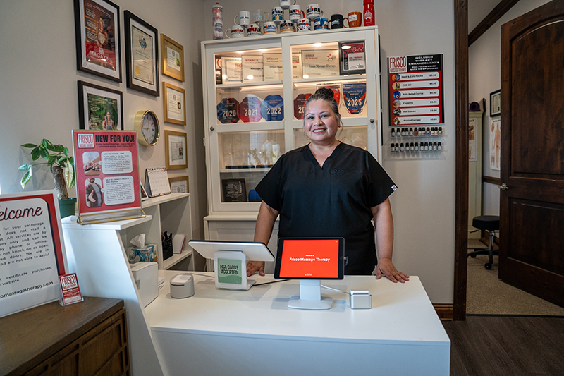 A woman in scrubs stands behind a reception desk in a wellness clinic, surrounded by awards, certificates, and wellness products that highlight the massage benefits and showcase the advantages of lymphatic drainage techniques.