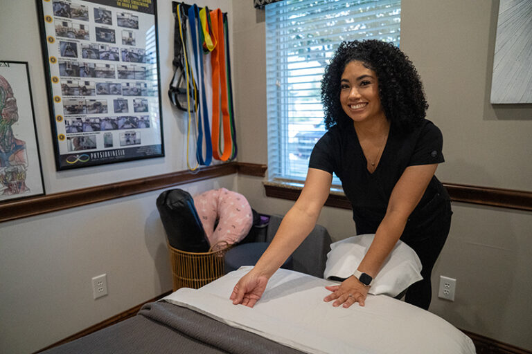 A woman in scrubs prepares a bed in a Frisco Massage Therapy room, smiling as she arranges pillows and linens. Therapy equipment and posters about the science behind sports therapy are visible in the background.