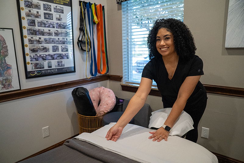 A woman in scrubs prepares a bed in a Frisco Massage Therapy room, smiling as she arranges pillows and linens. Therapy equipment and posters about the science behind sports therapy are visible in the background.