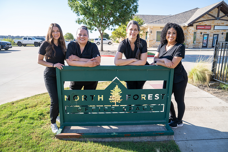 Four women in black outfits stand smiling behind a green "North Forest Office Space" sign outdoors on a sunny day, representing Craniosacral Therapy, with buildings and cars visible in the background.