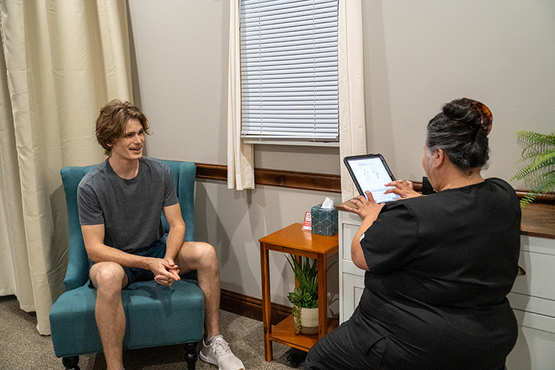 A man sits in a chair while a woman in scrubs enters information on a tablet in a small office with neutral decor, discussing the Top 5 benefits of craniosacral therapy for overall wellness.