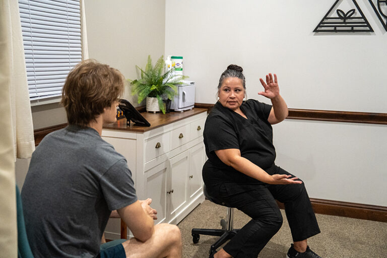 A woman in black scrubs gestures while discussing post-surgery therapy with a seated man in a casual shirt inside a small office or consultation room.