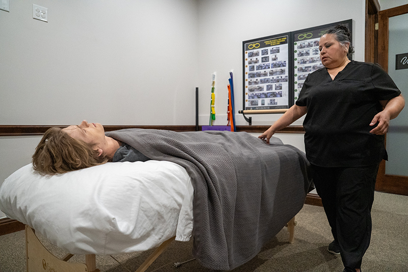 A person lies on a massage table covered with a gray blanket while a staff member in black scrubs stands nearby, offering a soothing lymphatic drainage session in the treatment room for pain-free living.