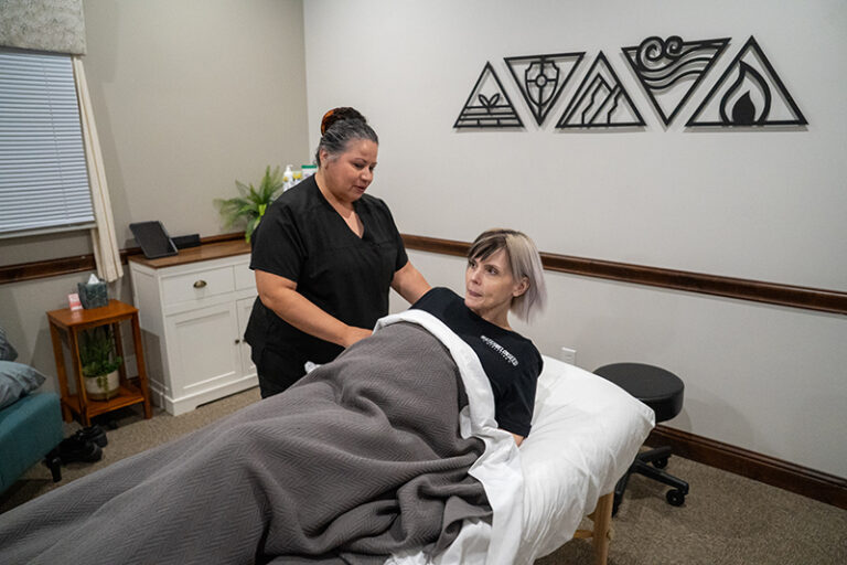 A woman enjoys a Prenatal Massage on a cozy table covered with a gray blanket while a therapist from Frisco Massage Therapy stands beside her in a treatment room adorned with geometric wall art.
