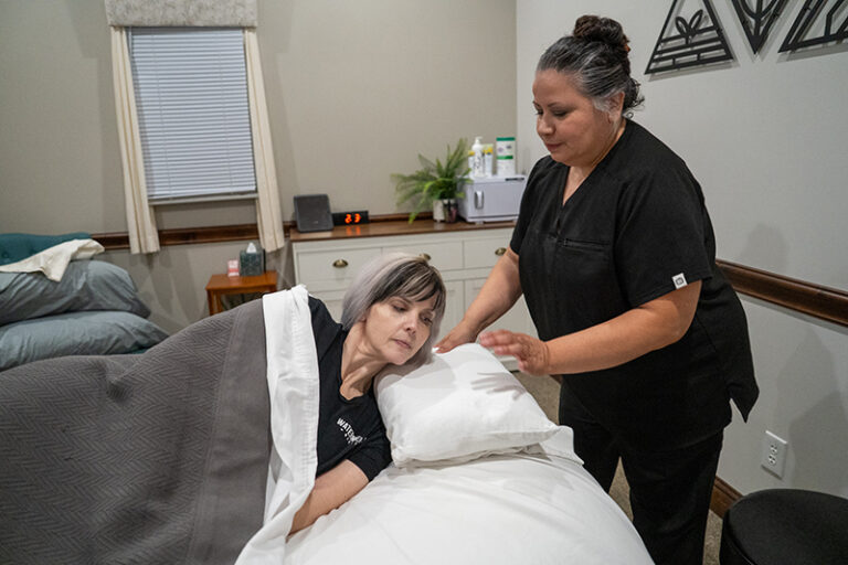 A woman lies in bed under a blanket while another woman in black scrubs, providing postnatal massage, adjusts her pillow in a well-lit room with neutral decor, supporting her recovery.