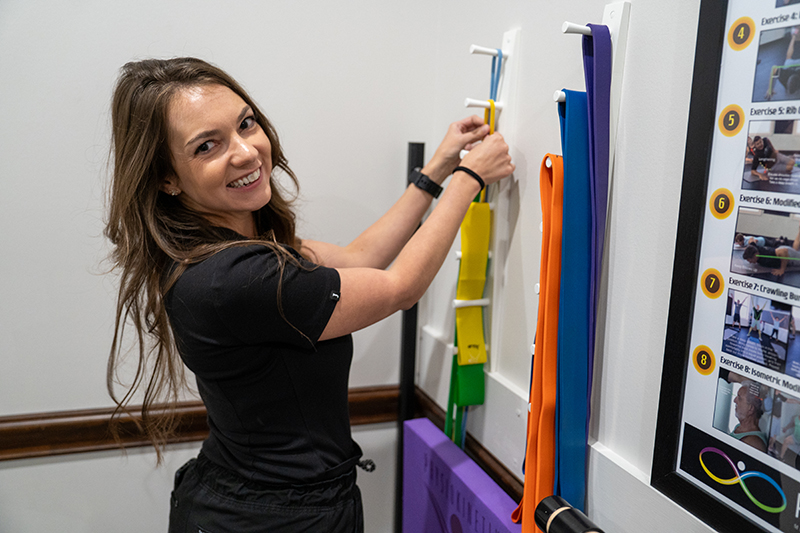 A woman smiles at the camera while organizing colorful resistance bands on a wall-mounted rack in a Sports Therapy or physical therapy setting, showing her commitment to mastering the game of recovery.