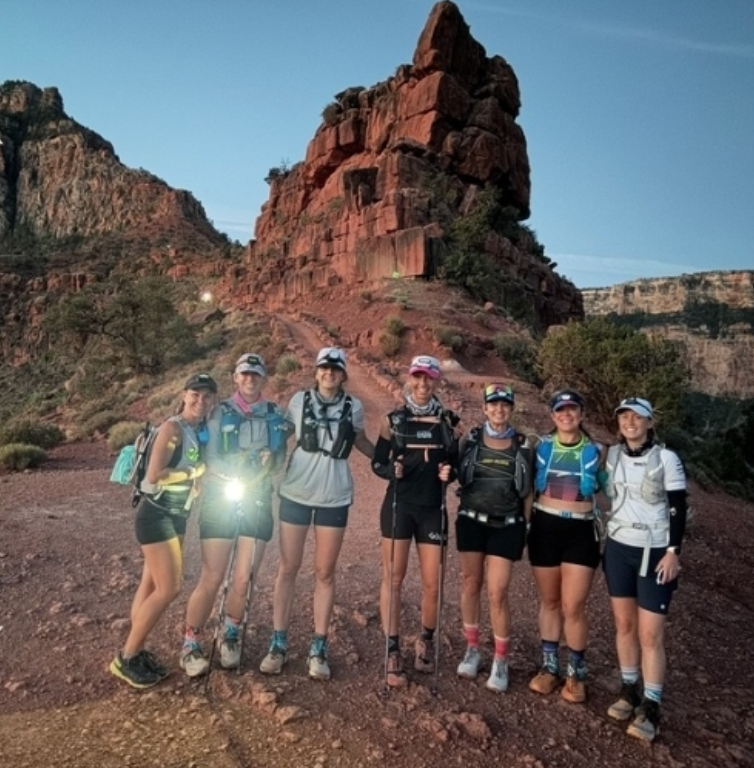 Seven women in running gear stand together on a rocky trail with a large red rock formation in the background at sunrise, embodying the harmony of movement and nature—truly music as medicine for the soul.