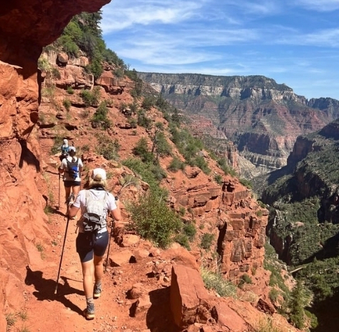 Two hikers with backpacks and trekking poles walk along a narrow, rocky trail on a red canyon cliff under a blue sky, as if letting the rhythm of nature serve as music as medicine for their souls.