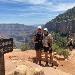 Two hikers pose on a rocky overlook with a canyon view behind them. A sign reading "ONINO OVERLOOK" is visible on the left, as they discuss how Music as Medicine inspires their outdoor adventures.
