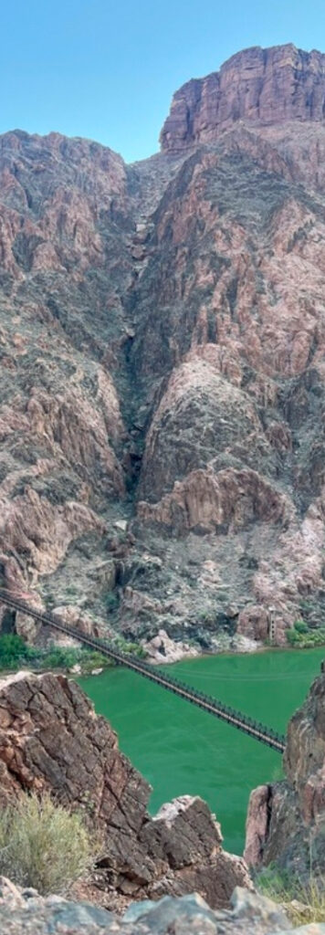 A suspension bridge spans a green river at the base of steep rocky cliffs under a clear blue sky, where the serene scene embodies the harmony and calm found in music as medicine.