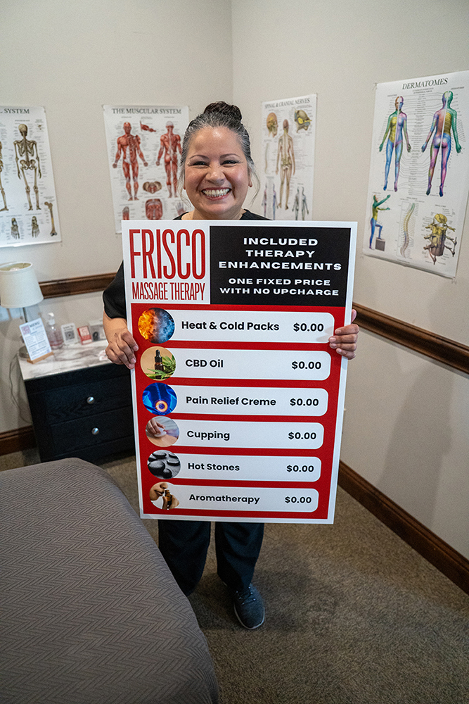 A smiling person stands in a therapy room holding a sign listing free massage therapy enhancements—perfect for sports therapy Frisco or injury recovery Frisco—including heat packs, CBD oil, pain creme, cupping, hot stones, and aromatherapy.