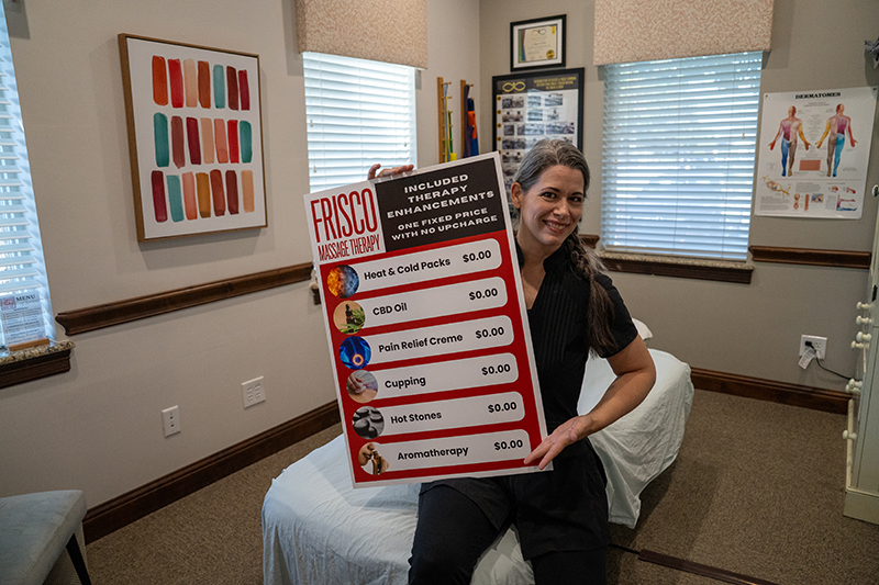 A woman sits on a massage table in a clinic room, holding a large sign listing free therapy enhancements at Frisco Massage Therapy, including TMJ massage Frisco for effective jaw pain relief.