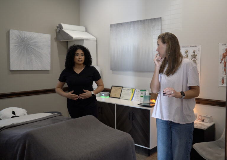 Two women stand in a medical or therapy room; one in scrubs gestures while the other listens with a hand near her mouth. A treatment bed and anatomical chart are visible.