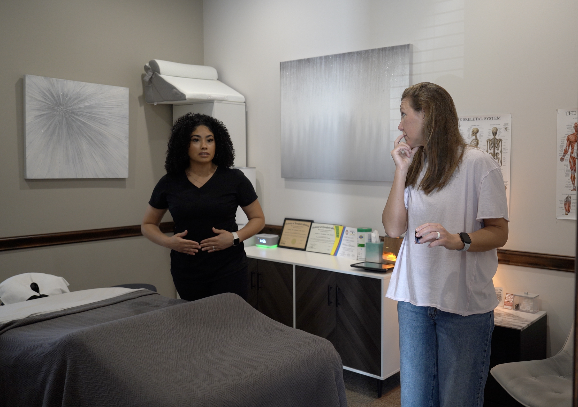 Two women stand in a medical or therapy room; one in scrubs gestures while the other listens with a hand near her mouth. A treatment bed and anatomical chart are visible.