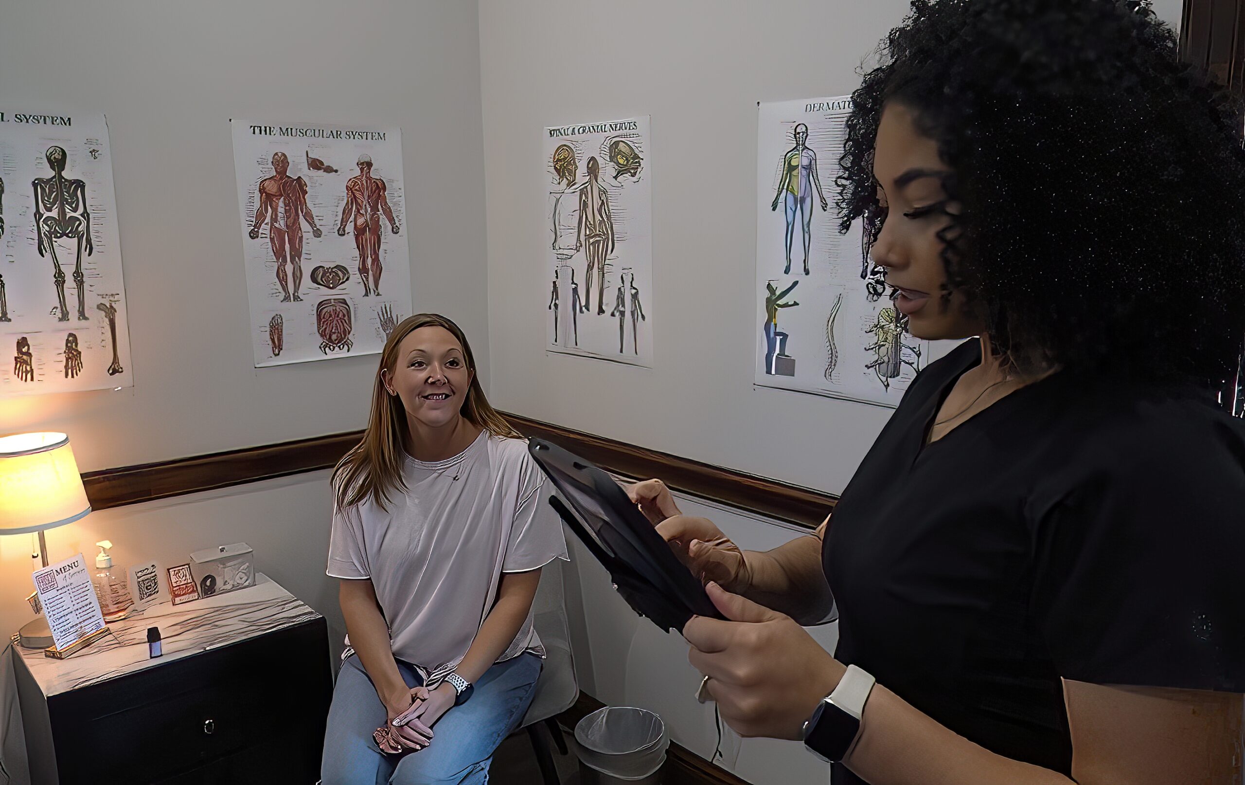 A woman sits on an exam table while a healthcare professional reviews Manual Lymphatic Drainage Techniques on a tablet in a medical office with anatomy posters on the walls.