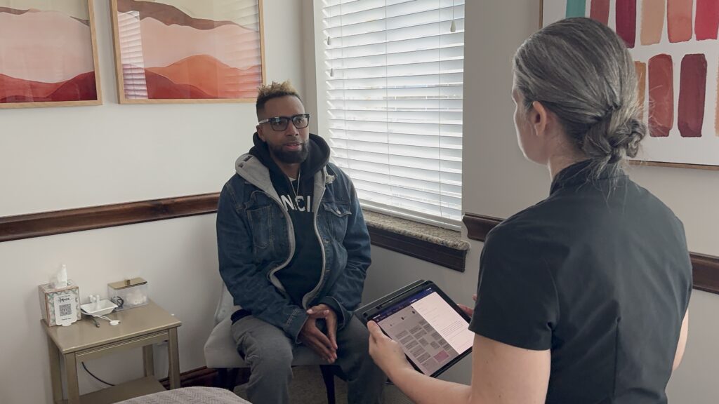 A person sits on a chair in a medical office while another person stands nearby holding a tablet, appearing to conduct an interview or consultation.