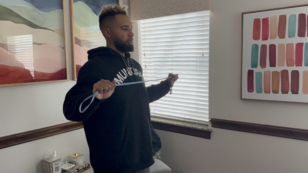 A man stands indoors wearing a black hoodie, holding and pulling a resistance band in front of a window with blinds and colorful artwork on the walls.
