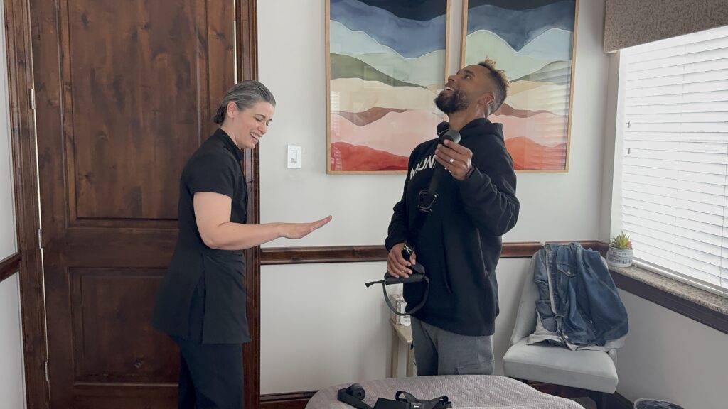 A woman and a man stand in a room; the woman gestures with her hand while the man, holding a headset, looks up and smiles. Casual clothes and a jacket are seen on a bed nearby.