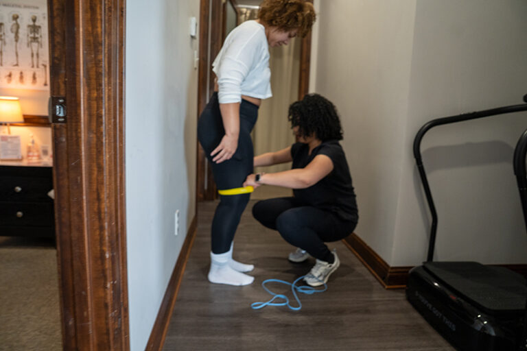 A person squats while another measures their thigh with a yellow tape measure in a hallway, next to exercise equipment.
