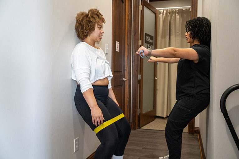 A woman performs a wall sit with a resistance band around her thighs while an instructor observes and checks a device, possibly a timer, in a hallway.