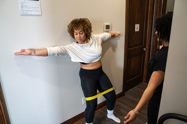 A woman stands against a wall with a resistance band around her thighs, arms outstretched, while another person observes her in a room.