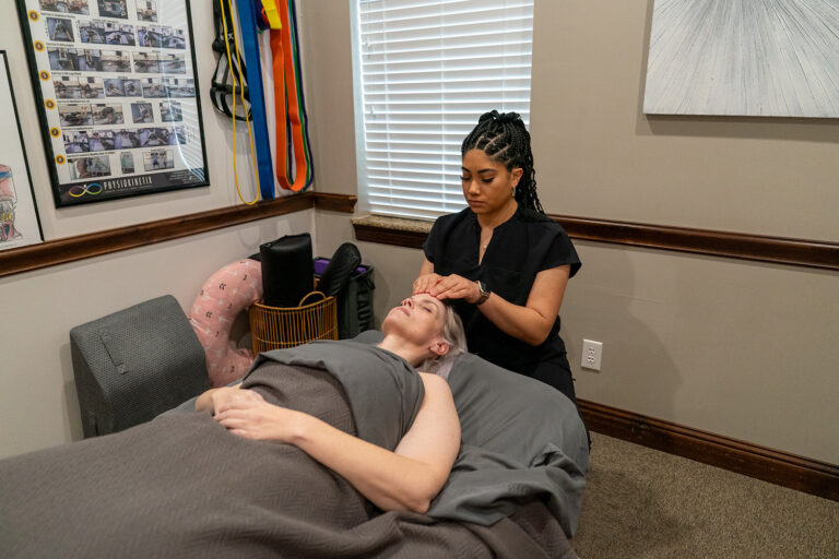 A massage therapist performs a facial massage on a client lying on a massage table in a treatment room.