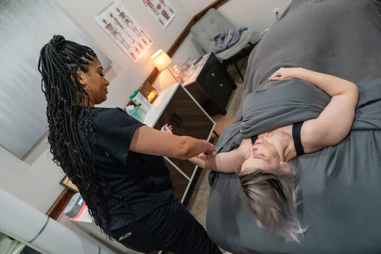 A massage therapist works on the arm of a woman lying on a massage table, partially covered with a gray blanket in a treatment room.