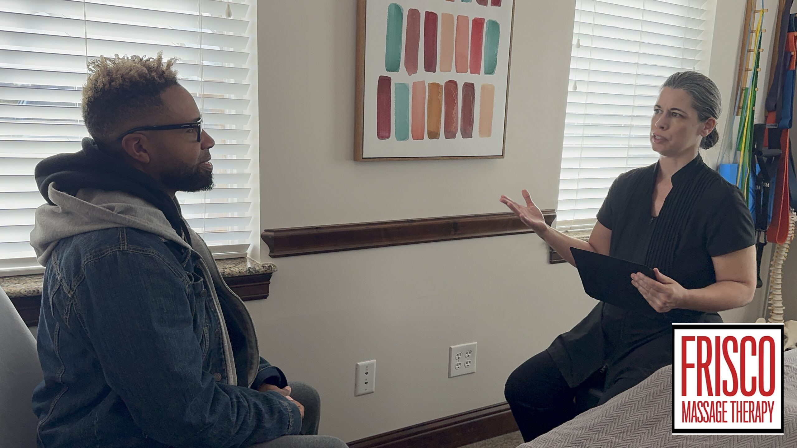 A man sits across from a woman holding a clipboard in a consultation room at Frisco Massage Therapy, discussing therapeutic massage for athletes.