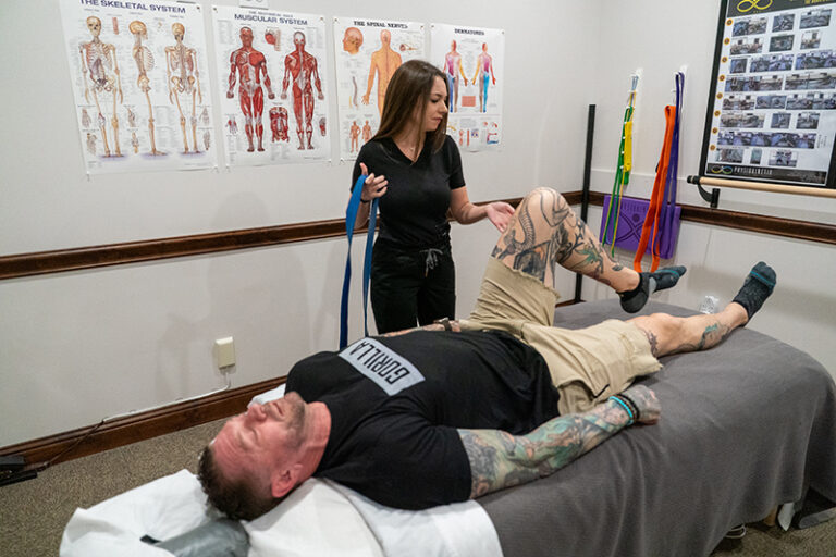 A physical therapist assists a man lying on a treatment table with a leg exercise in a clinic, where anatomical charts highlight the benefits of therapeutic massage for athletes.