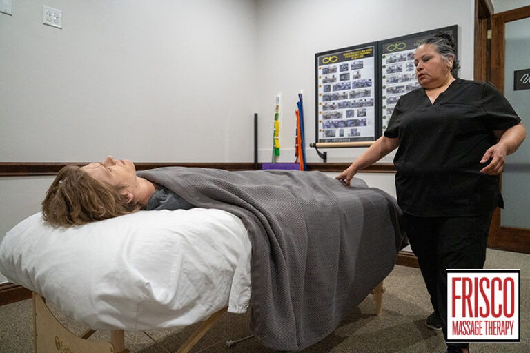 A person lies on a massage table covered with a blanket while a therapist stands nearby in a clinic room, offering therapeutic massage Frisco style. The "Frisco Massage Therapy" logo is visible in the lower right corner.