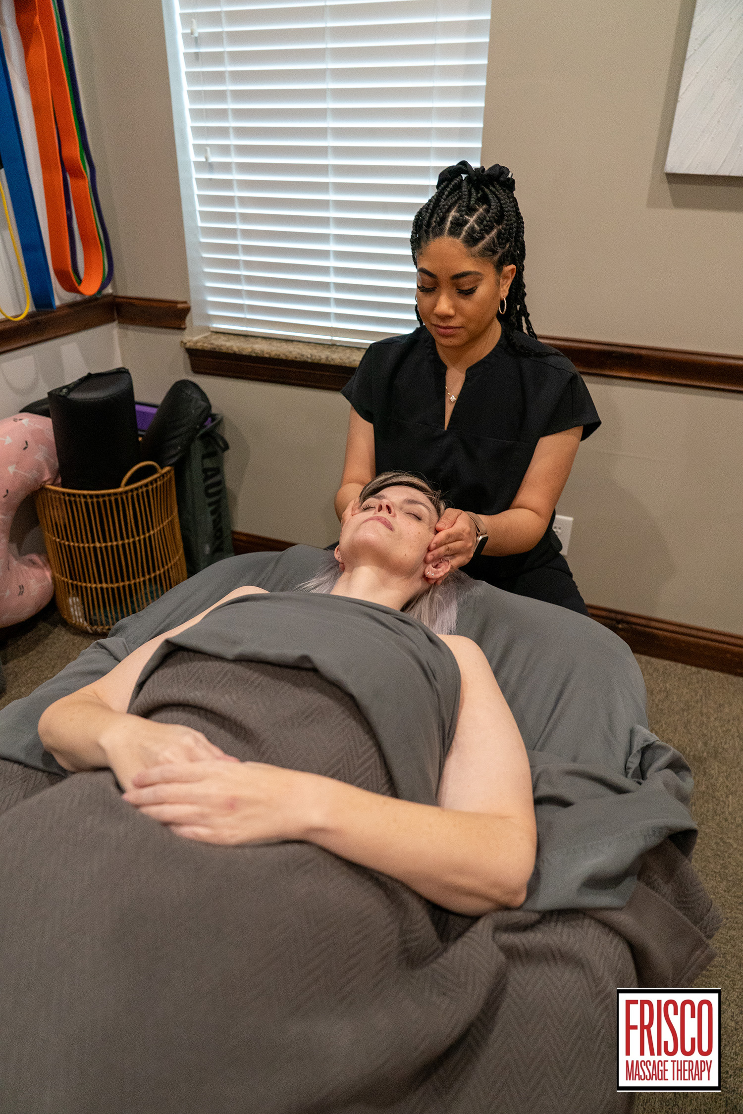 A massage therapist gives a facial massage to a person lying on a table under a blanket in a treatment room, highlighting Frisco Massage Therapy’s personalized massage therapy plans. The logo is visible in the corner.
