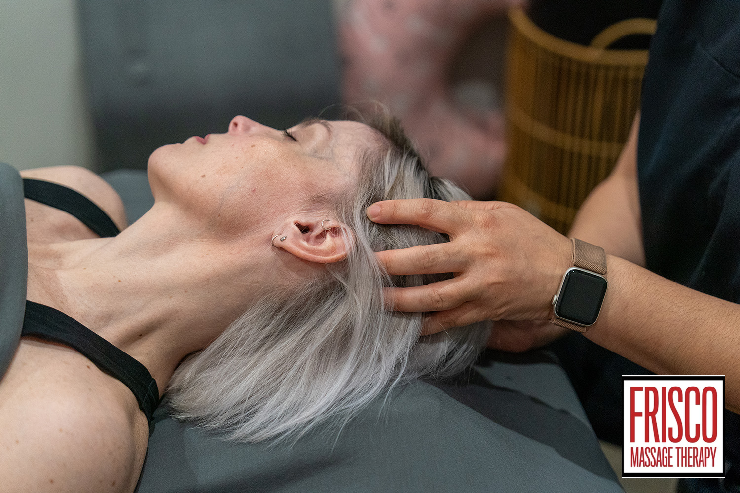 A person with gray hair receives a head massage from a therapist, known for being lymphatic drainage specialists. The Frisco Massage Therapy logo is visible in the lower right corner.