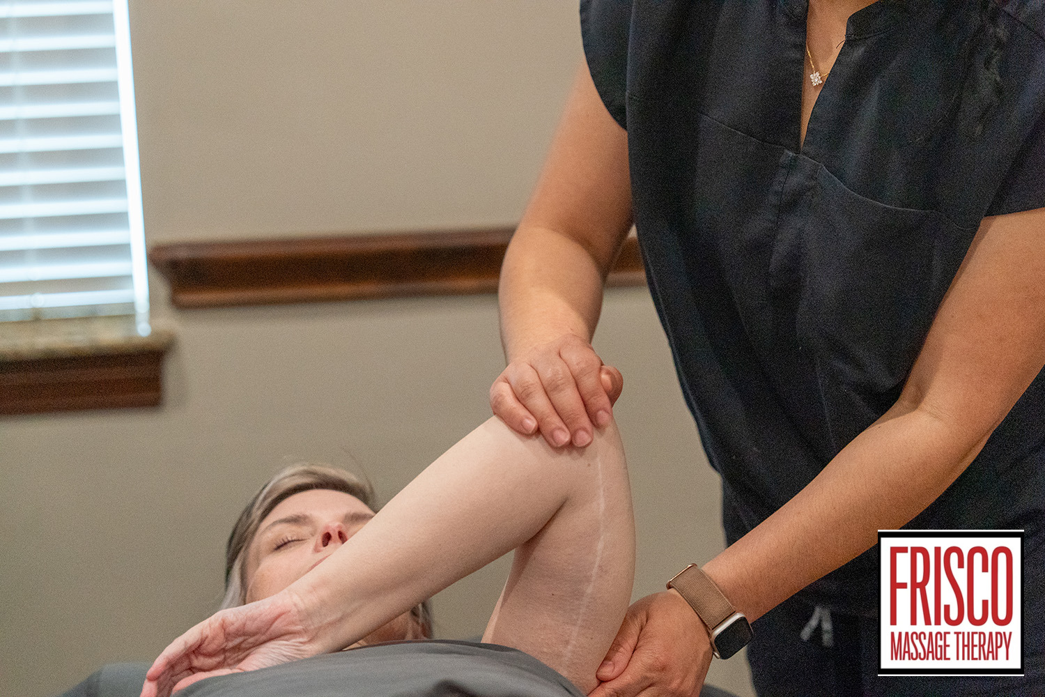A massage therapist performs manual lymphatic drainage by gently stretching a client's arm during a session. The client is lying down with eyes closed. "Frisco Massage Therapy" logo is visible in the corner.