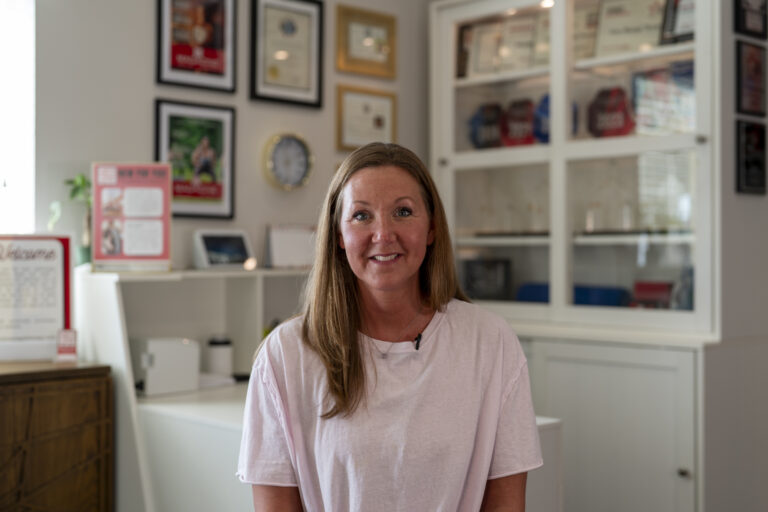 A woman with long hair wearing a light pink shirt sits and smiles in an office with framed certificates and awards on the wall behind her.