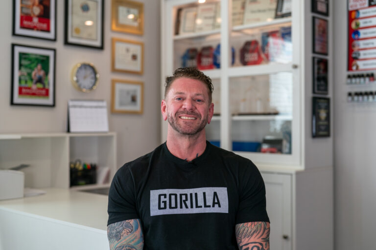 A man with tattoos wearing a black "GORILLA" t-shirt sits and smiles in a Frisco office with awards, certificates, and a clock on the wall behind him—ready to share his athlete testimonial about therapeutic massage.
