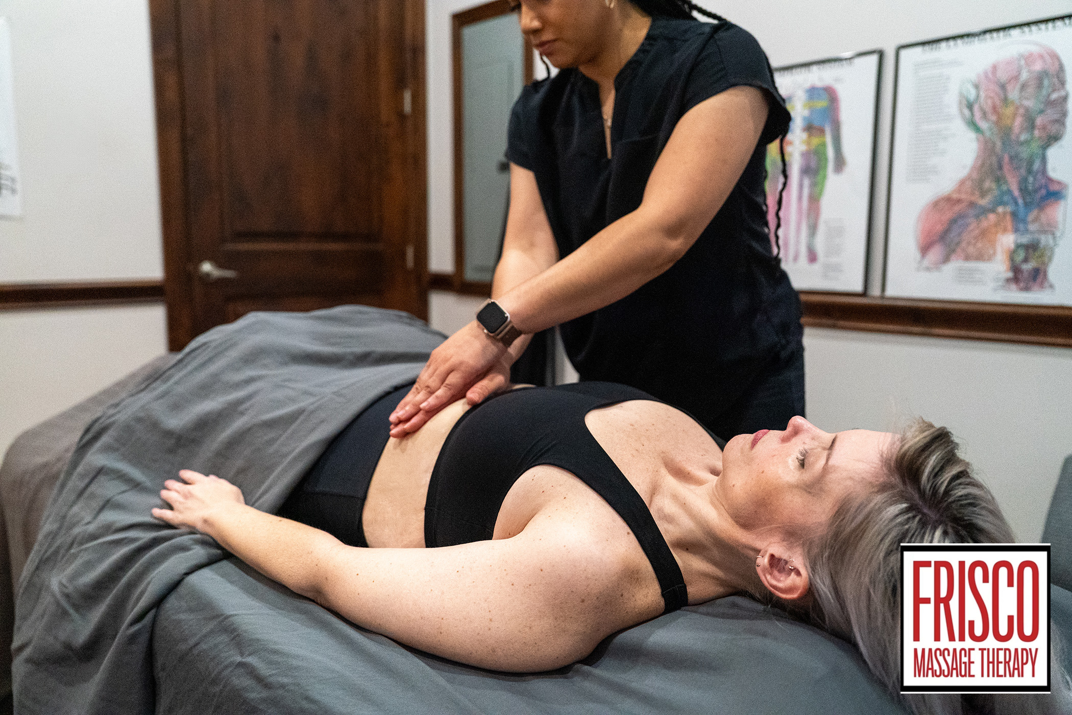 A massage therapist performs abdominal massage for scar pain on a woman lying on a table in a clinic room. The "Frisco Massage Therapy" logo is visible in the lower right corner.