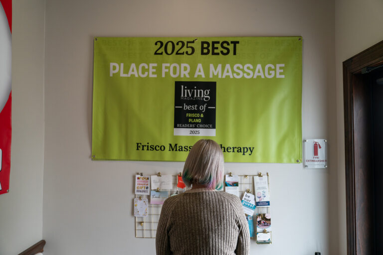 A person with gray hair faces a green banner that reads "2025 Best Place for a Massage, Frisco Massage Therapy," displayed above a wall of brochures and cards.