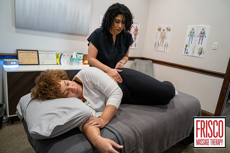 A massage therapist performs a side-lying hip massage on a client in a clinic, with anatomical charts and certificates on the walls, demonstrating how prenatal massage helps pregnancy by providing comfort and support.